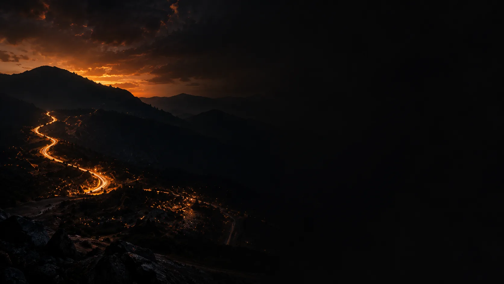 Dark mountain valley at dusk with a winding road lit by golden car-light trails, under a sunset sky
